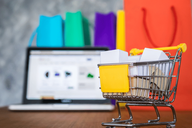 Carrinho de compras em miniatura com caixas de papelão em frente a um laptop e sacolas coloridas ao fundo, simbolizando compras online e vendas pela internet.