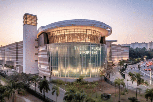 Imagem do Tietê Plaza Shopping, um centro de compras moderno com fachada de vidro e iluminação, localizado em uma área urbana com árvores e movimento de carros ao redor.