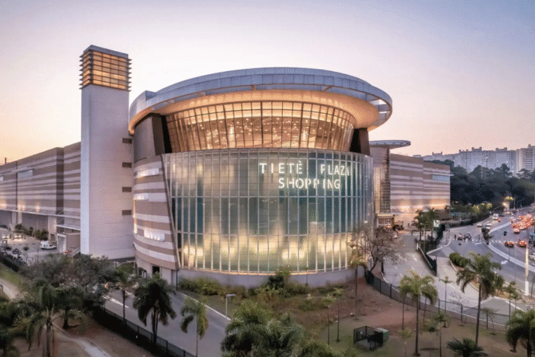 Imagem do Tietê Plaza Shopping, um centro de compras moderno com fachada de vidro e iluminação, localizado em uma área urbana com árvores e movimento de carros ao redor.