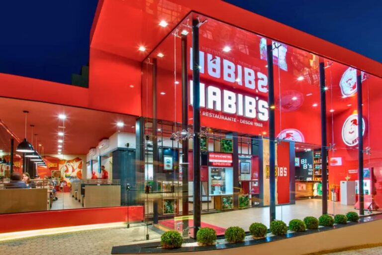 Fachada iluminada de um restaurante Habib’s com interior moderno e clientes sentados nas mesas.