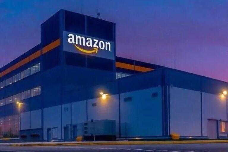 Imagem de um centro de distribuição da Amazon à noite, com luzes acesas e logo visível na fachada do prédio.