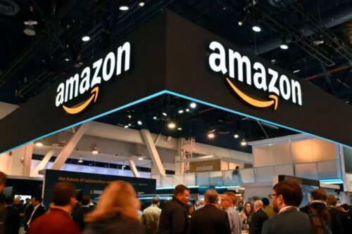 Imagem do estande da Amazon na feira de tecnologia com diversas pessoas ao redor, destaque para o logo da Amazon no teto, em ambiente de evento corporativo.