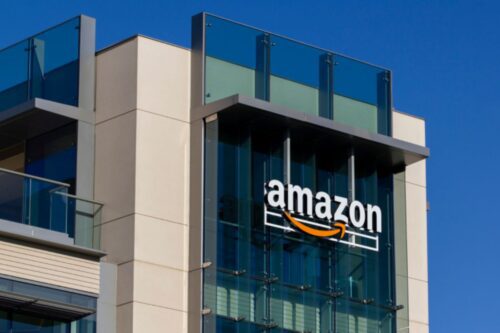 Imagem moderna da fachada do prédio da Amazon com vidro e estrutura em tons claros, mostrando a logo da Amazon na parte superior de um edifício de escritórios, sob um céu azul claro.