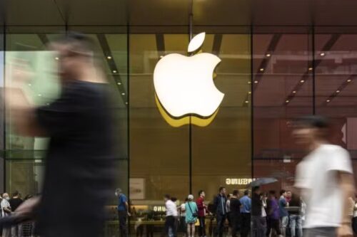 Fachada de uma loja da Apple com grande logotipo iluminado e várias pessoas passando em frente, destacando a presença de consumidores e movimento na área.