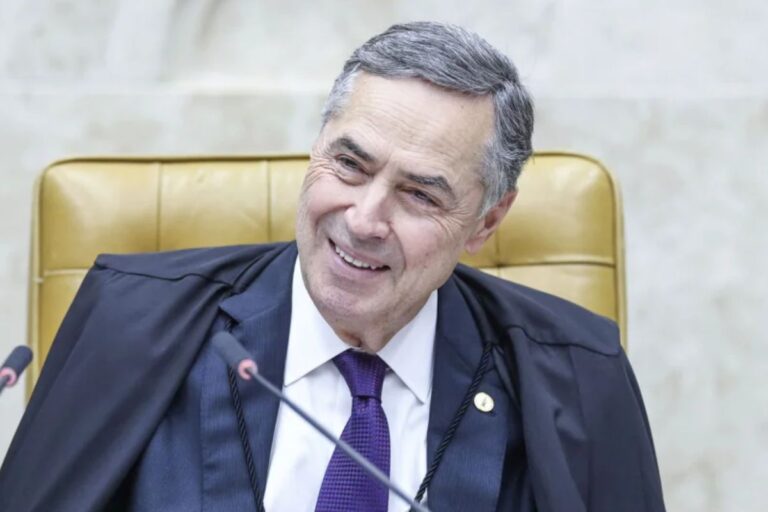 Barroso sorridente usando toga preta, camisa branca e gravata roxa, sentado em uma cadeira de tribunal