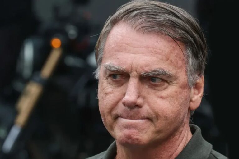 Imagem de Bolsonaro com expressão séria, cabelo grisalho penteado para trás, pele com sinais de envelhecimento e um fundo escuro desfocado.