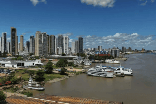 Paisagem urbana de Belém com arranha-céus altos ao lado de um rio, com vários barcos atracados, sob céu azul com nuvens, em uma cidade cosmopolita.