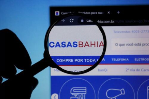 Imagem de uma pessoa segurando uma lupa que destaca o logo do site Casas Bahia na tela de um computador, promovendo compras online de móveis, eletrônicos e eletrodomésticos.