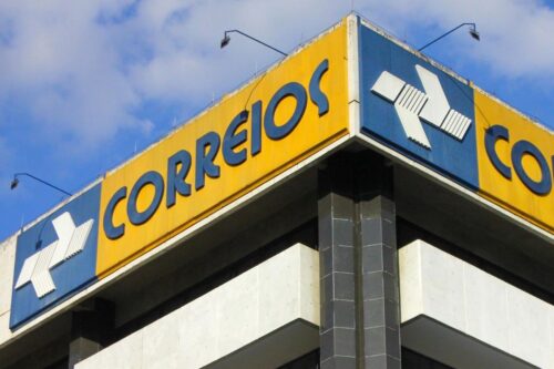 Imagem da fachada de uma agência dos Correios com identidade visual amarela, azul e o logo característico em destaque, localizado em um edifício alto com céu azul ao fundo.