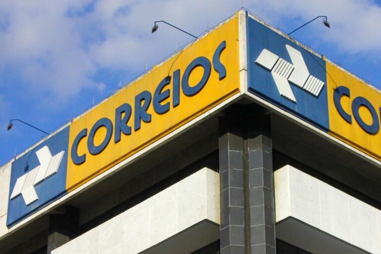 Imagem da fachada de uma agência dos Correios com identidade visual amarela, azul e o logo característico em destaque, localizado em um edifício alto com céu azul ao fundo.