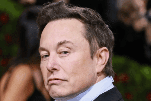 Elon Musk com expressão séria, cabelo castanho escuro e pele clara, usando terno preto e cabelo bem ajustado, em fundo desfocado.