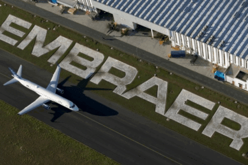 Avião decolando ao lado do grande LOGO da Embraer no chão de um aeroporto, destaque para o nome da fabricante brasileira de aeronaves.