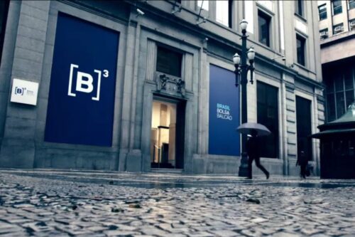 Fachada de um edifício de escritórios com sinais de branding do Brasil Bolsa Balcão (B3), localizado na cidade, com uma pessoa caminhando com guarda-chuva em um dia chuvoso.