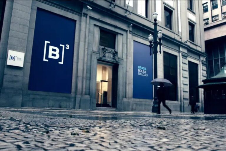 Fachada de um edifício de escritórios com sinais de branding do Brasil Bolsa Balcão (B3), localizado na cidade, com uma pessoa caminhando com guarda-chuva em um dia chuvoso.