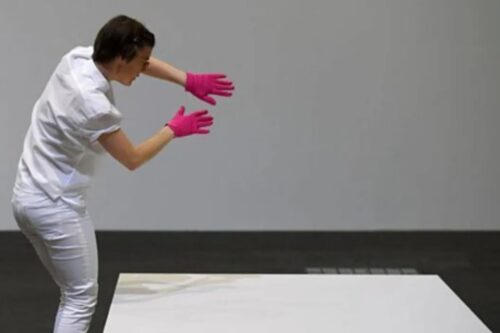 Pessoa "manipulando" a escultura invisível usando os braços e com expressão de esforço, vista de lado, no interior de um ambiente neutro.