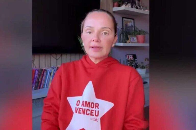 "Esquerdogata" usando blusa vermelha com estampa de estrela e escrita 'O amor venceu', em ambiente com livros e decoração de casa ao fundo.