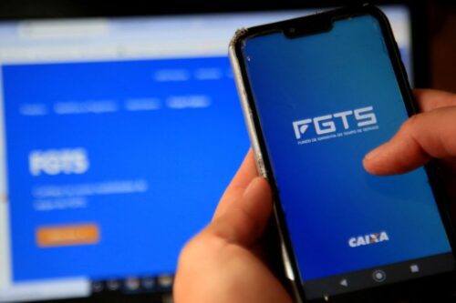 Pessoa utilizando o aplicativo do FGTS no celular, em frente a uma tela de computador com informações sobre o programa.