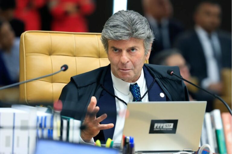 Imagem de Fux, sentado em uma cadeira com expressão séria, usando toga e frente a uma mesa com documentos e computador.