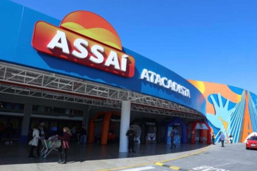 Entrada do mercado Assaí Atacadista com fachada azul e logotipo vermelho e amarelo sob céu claro, com pessoas entrando e saindo do local.