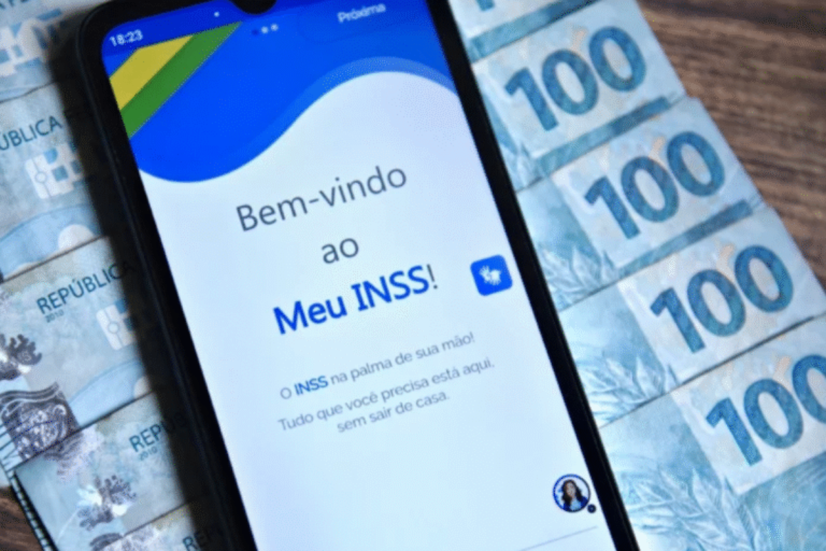 Imagem de um celular exibindo a tela de login do INSS com uma mensagem de boas-vindas, ao lado há várias notas de cem reais.