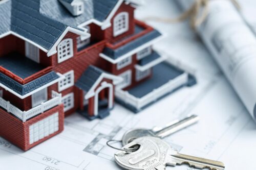 Maquete de uma casa de design detalhado, com chaves de metal no primeiro plano, simulando planejamento e financiamento imobiliário, sobre um projeto arquitetônico.