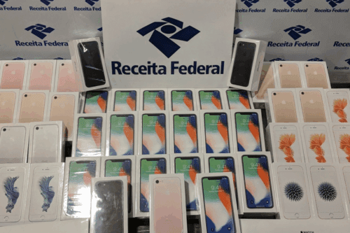 Diversos aparelhos de celular, incluindo iPhones e outros modelos, exibidos em uma mesa na presença de um banner da Receita Federal, indicando provável apreensão ou fiscalização.
