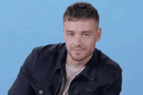 Liam Payne com cabelo curto e barba, usando jaqueta de tricô e camiseta, sentado contra fundo azul claro, sorrindo levemente para a câmera.