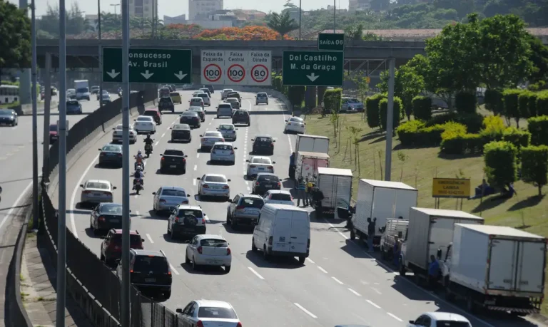 Foto mostra a Linha Amarela, via expressa que liga a Ilha do Fundão à Barra da Tijuca, no Rio de Janeiro, com intenso fluxo de veículos sob céu claro.