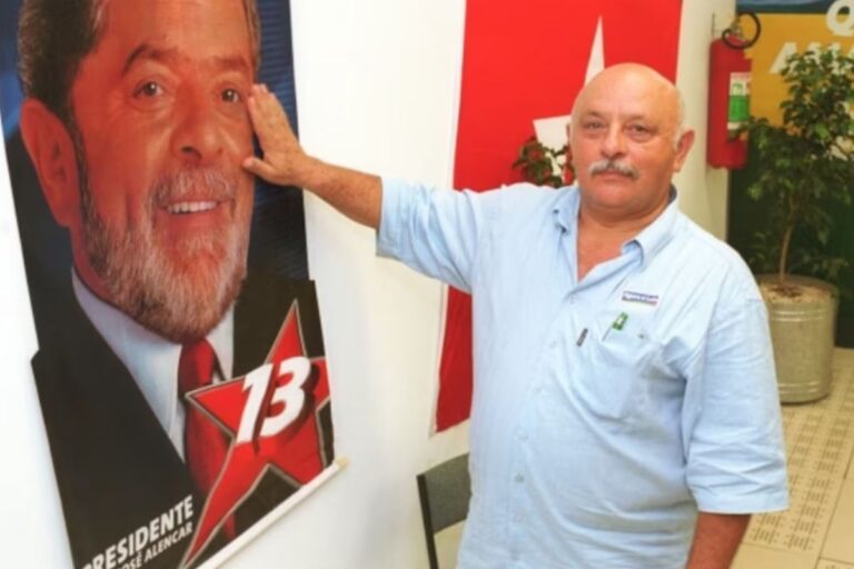 Irmão de Lula tocando a cabeça de um pôster de campanha eleitoral de um candidato político, na parede de um ambiente interno.