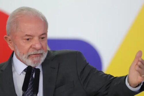 Lula de cabelos brancos e barba, vestindo terno escuro, fazendo uma apresentação ou discurso em um evento, com fundo colorido e iluminado.