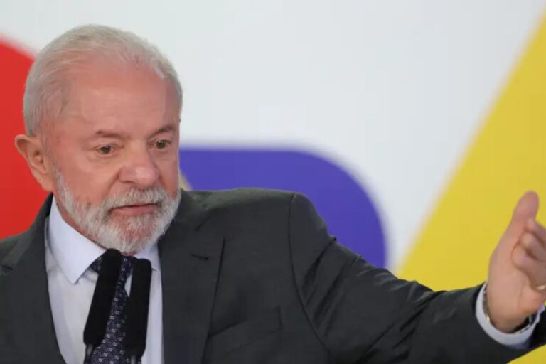 Lula de cabelos brancos e barba, vestindo terno escuro, fazendo uma apresentação ou discurso em um evento, com fundo colorido e iluminado.