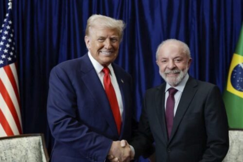 Reunião entre Donald Trump e Luiz Inácio Lula da Silva em evento político, com ambos sorrindo e apertando as mãos, ao fundo bandeira dos Estados Unidos e do Brasil.