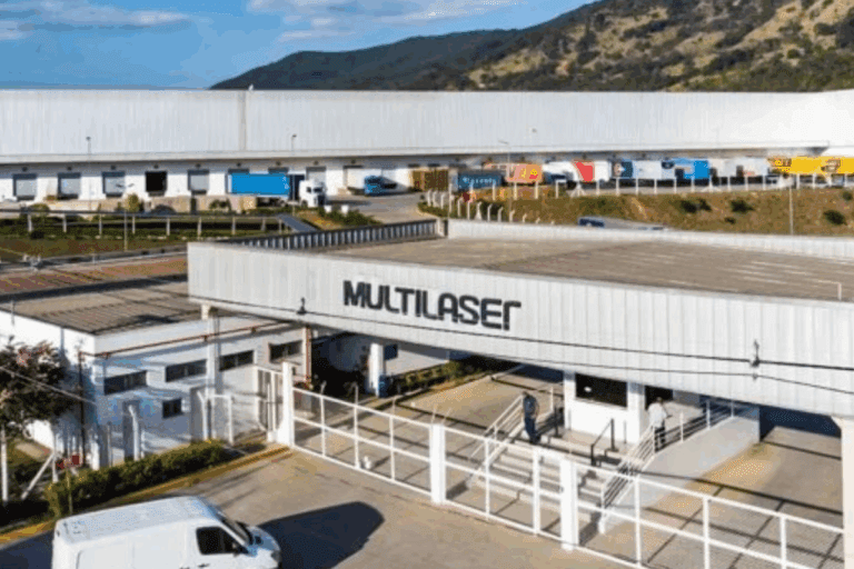Imagem do centro de distribuição Multlaser com fila de caminhões carregados na frente, localizado em uma região com relevo montanhoso ao fundo, sob céu claro.