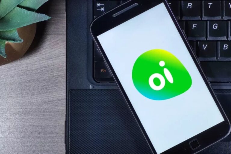 Smartphone com o aplicativo oi na tela, descansando sobre um teclado de computador preto sob uma mesa de madeira clara, ao lado de uma planta. Ideal para quem busca informações sobre planos de celular e internet móvel.