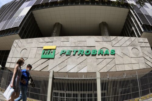 Imagem da sede da Petrobras com logo verde e pessoas caminhando na frente do prédio em horário diurno