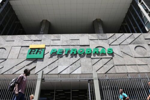 Fachada do edifício da Petrobras com logo em destaque, com pessoas caminhando na frente em um dia ensolarado.