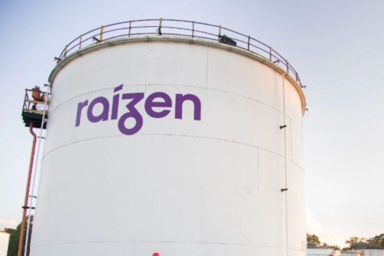 Tanque de armazenamento de produtos com a marca Raízen escrita em roxo, localizado ao ar livre sob céu claro, símbolo de energia e distribuição de combustíveis.