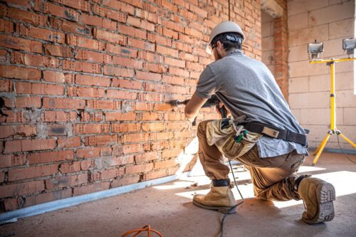 Empreiteiro trabalhando na construção civil, instalando uma parede de tijolos com segurança usando capacete e ferramentas, dentro de um edifício em construção.
