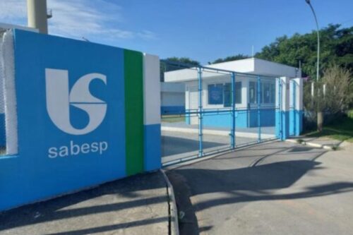 Entrada principal do posto de atendimento da Sabesp com portão azul, sinalização e entorno de área verde, em dia de sol com céu claro.