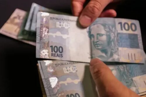 Pessoa segurando notas de 100 reais, dinheiro em cédulas brasileiras, com destaque para as características das notas.