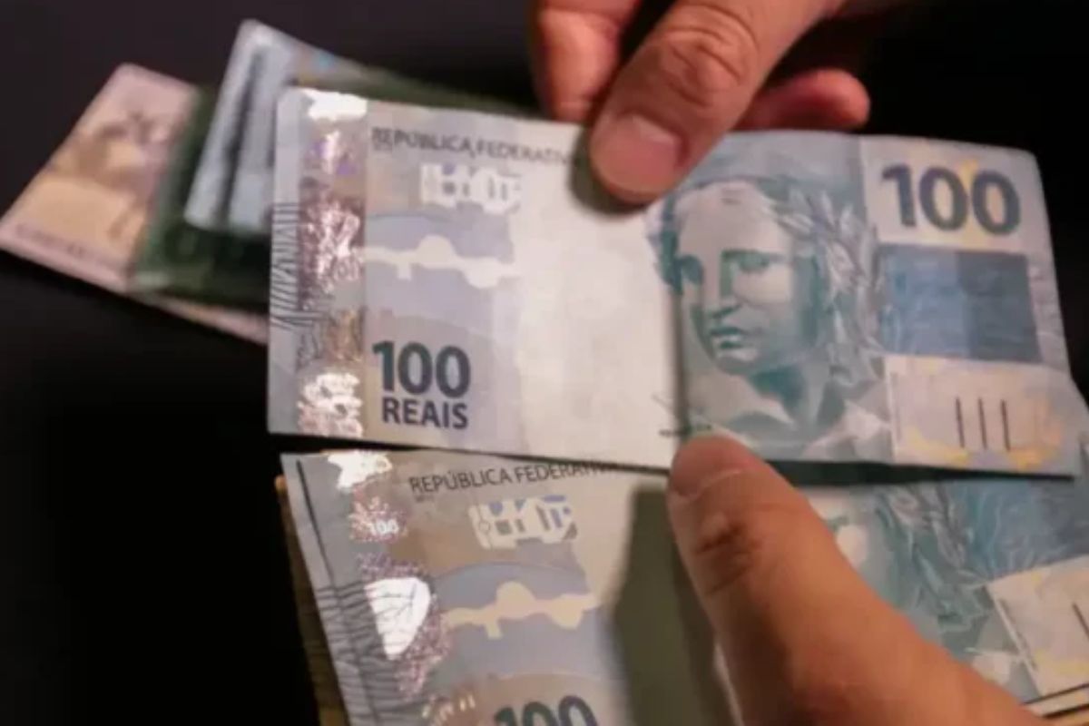 Pessoa segurando notas de 100 reais, dinheiro em cédulas brasileiras, com destaque para as características das notas.