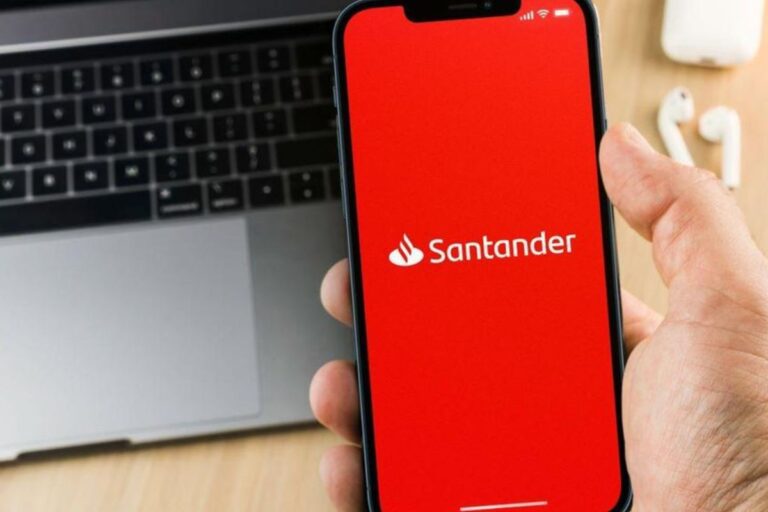 Pessoa segurando um smartphone exibindo o aplicativo do banco Santander, com um teclado de laptop ao fundo.