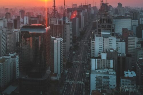 Vista de um skyline urbano ao pôr do sol, com prédios altos e ruas movimentadas, na cidade de São Paulo ao entardecer.
