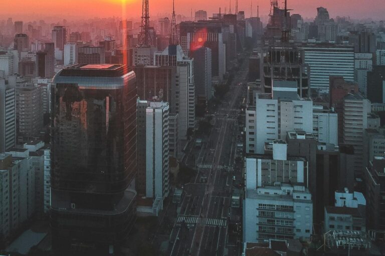 Vista de um skyline urbano ao pôr do sol, com prédios altos e ruas movimentadas, na cidade de São Paulo ao entardecer.