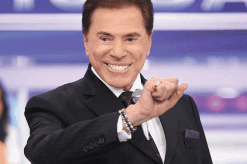 Silvio Santos sorridente segurando microfone, vestindo terno preto e gravata no palco, celebrando sucesso na televisão brasileira.