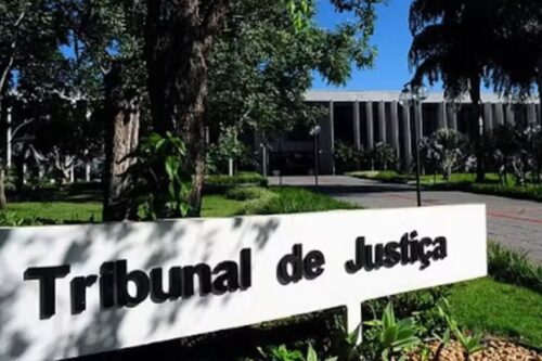 Fachada do Tribunal de Justiça ao ar livre com um painel branco e árvores verdes ao fundo, sob céu azul sólido.