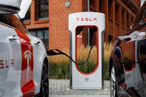 Carros elétricos Tesla conectados à estação de carregamento em frente a um prédio de tijolos, evidenciando infraestrutura de veículos elétricos moderna.