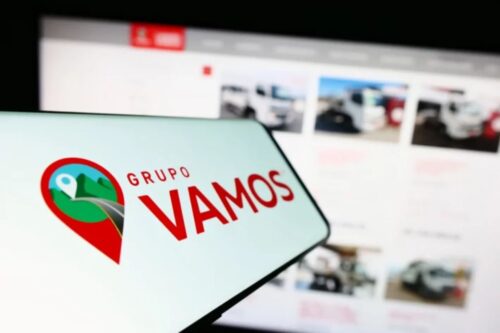 Imagem de um smartphone com o logo do Grupo VAMOS, uma empresa de localização por GPS, com uma tela ao fundo mostrando um site de vendas de carros.