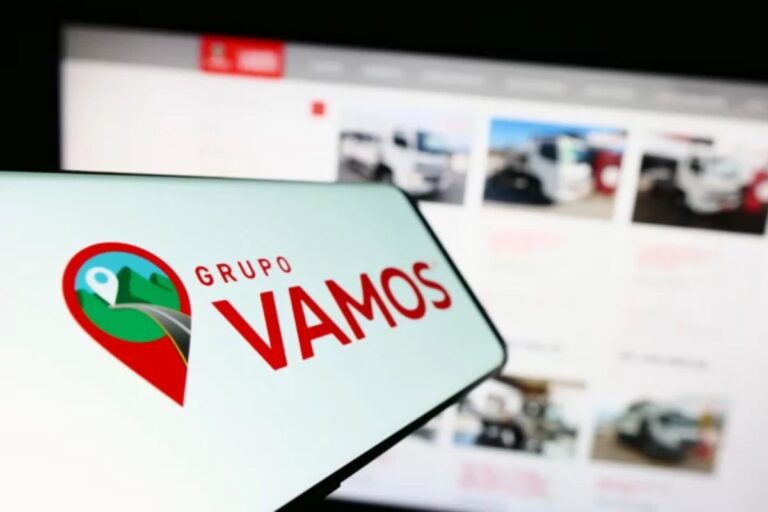 Imagem de um smartphone com o logo do Grupo VAMOS, uma empresa de localização por GPS, com uma tela ao fundo mostrando um site de vendas de carros.