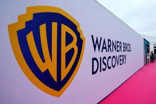 Logotipo da Warner Bros. Discovery em uma parede branca em um evento, com destaque para o símbolo clássico da Warner Bros. e o nome da empresa ao lado.